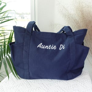 May include: A navy blue tote bag with two side pockets and sturdy handles. The words "Auntie Di" are embroidered in white script across the front. The bag is made of a durable fabric and is designed for carrying items.