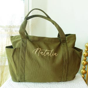 May include: An olive green tote bag with two exterior pockets and sturdy handles. The name "Natalia" is embroidered in a cursive font on the front of the bag. The bag is made of a durable canvas material.