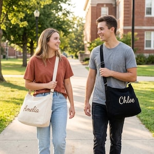 May include: Two people walking on a path, each carrying a personalized canvas messenger bag. One bag is off-white with "Amelia" embroidered, the other is black with "Chloe". They are wearing casual clothes.