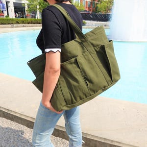 May include: An olive green canvas tote bag with multiple exterior pockets. The bag has a large main compartment and a shoulder strap. The person is wearing a black shirt and blue jeans. The background includes a fountain and buildings.