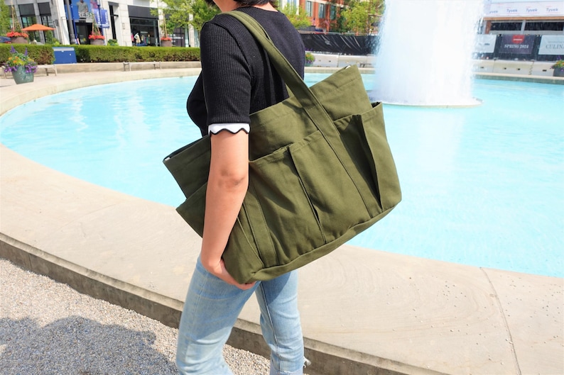 May include: An olive green canvas tote bag with multiple exterior pockets. The bag has a large main compartment and a shoulder strap. The person is wearing a black shirt and blue jeans. The background includes a fountain and buildings.