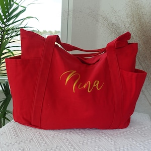 May include: A red tote bag with two exterior pockets and long handles. The word "Nina" is embroidered in gold script on the front of the bag. The bag is made of a sturdy fabric and is sitting on a white surface.
