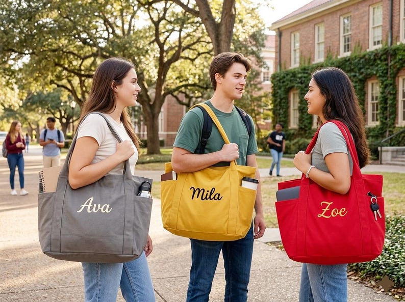 May include: Three students stand on a college campus, each carrying a personalized tote bag. The bags are gray, yellow, and red, with the names "Ava", "Mila", and "Zoe" embroidered on them. The scene suggests a back-to-school theme.