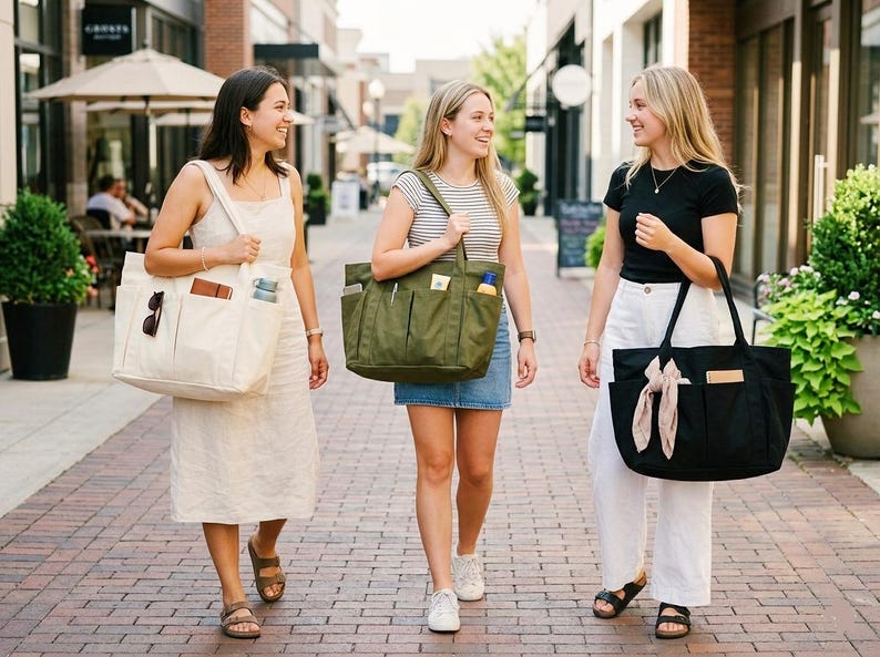May include: Three women walking outdoors, each carrying a tote bag. One bag is off-white, one is olive green, and the other is black. The bags have multiple pockets and are filled with various items. The women are smiling and appear to be enjoying a day out.