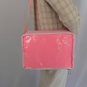K&ouml;nnte beinhalten: Eine rosa Rei&szlig;verschlusstasche mit einem transparenten Plastikfenster. Die Tasche hat einen Schultergurt und ist aus einem weichen, strapazierf&auml;higen Material gefertigt.