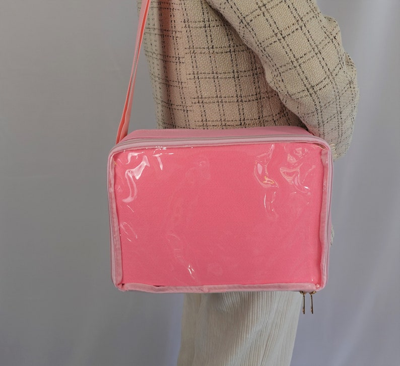 K&ouml;nnte beinhalten: Eine rosa Rei&szlig;verschlusstasche mit einem transparenten Plastikfenster. Die Tasche hat einen Schultergurt und ist aus einem weichen, strapazierf&auml;higen Material gefertigt.