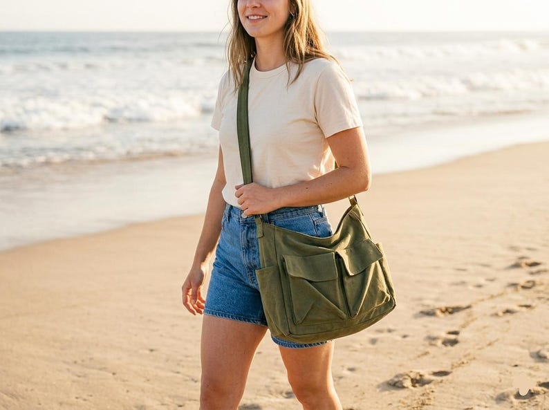 May include: An olive green canvas shoulder bag with multiple front pockets and a long strap. The bag is carried by a person walking on a sandy beach. The person is wearing denim shorts and a cream-colored t-shirt.