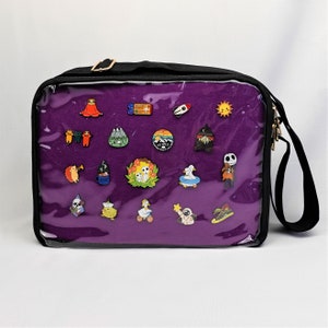 K&ouml;nnte beinhalten: Eine violette Rei&szlig;verschlusstasche mit einer durchsichtigen Vorderseite. Die Tasche hat einen schwarzen Riemen und ist mit verschiedenen Emaille-Pins gef&uuml;llt. Die Pins zeigen eine Vielzahl von Designs, darunter Tiere, Pflanzen und abstrakte Formen.