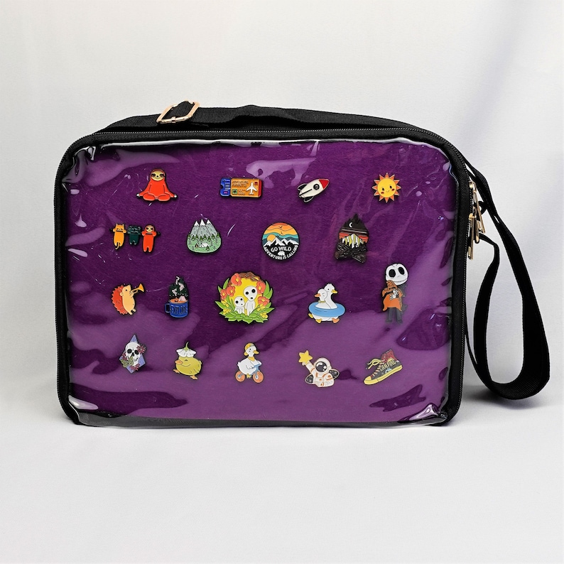 K&ouml;nnte beinhalten: Eine violette Rei&szlig;verschlusstasche mit einer durchsichtigen Vorderseite. Die Tasche hat einen schwarzen Riemen und ist mit verschiedenen Emaille-Pins gef&uuml;llt. Die Pins zeigen eine Vielzahl von Designs, darunter Tiere, Pflanzen und abstrakte Formen.