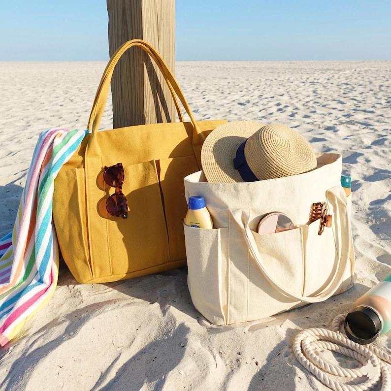 May include: Two canvas tote bags on a sandy beach. One is mustard yellow with sunglasses attached, the other is off-white and contains a straw hat, sunscreen, and a compact mirror. A striped towel rests nearby.