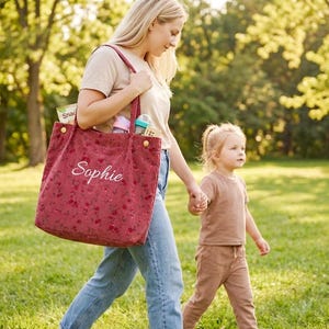 May include: A red tote bag with a floral pattern and the name "Sophie" embroidered in white. The bag is carried by a person walking with a child in a park. The bag contains snacks and baby supplies.