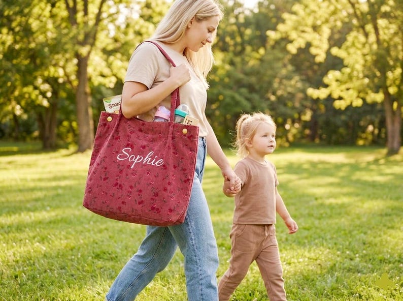 May include: A red tote bag with a floral pattern and the name "Sophie" embroidered in white. The bag is carried by a person walking with a child in a park. The bag contains snacks and baby supplies.
