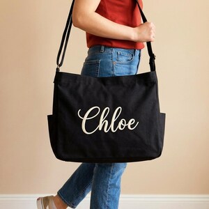 May include: Black canvas tote bag with the name "Chloe" in white script. The bag has a long shoulder strap and two exterior pockets. A casual and practical accessory.