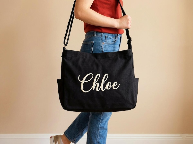 May include: Black canvas tote bag with the name "Chloe" in white script. The bag has a long shoulder strap and two exterior pockets. A casual and practical accessory.
