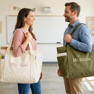 May include: Two canvas tote bags, one cream and one olive green, are held by a woman and a man. The cream bag has "Mrs. Taylor" in cursive, and the green bag has "Mr. Adams". Both bags have multiple pockets.