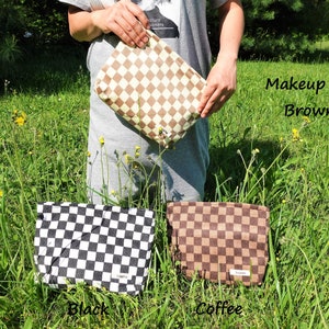 K&ouml;nnte beinhalten: Eine braune Make-up-Tasche mit Rautenmuster wird vor einem grasbewachsenen Hintergrund gehalten. Der Text "Makeup Bag Brown" ist sichtbar. Zwei weitere Taschen, eine schwarze und eine kaffeefarbene, werden darunter angezeigt.