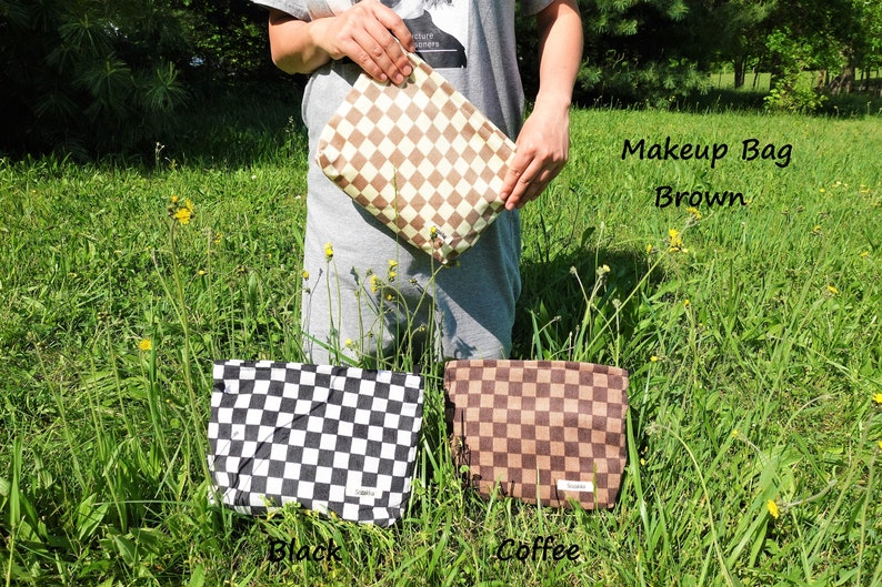 K&ouml;nnte beinhalten: Eine braune Make-up-Tasche mit Rautenmuster wird vor einem grasbewachsenen Hintergrund gehalten. Der Text "Makeup Bag Brown" ist sichtbar. Zwei weitere Taschen, eine schwarze und eine kaffeefarbene, werden darunter angezeigt.