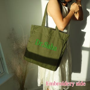 Pode incluir: Bolsa tote verde-oliva com o texto bordado "Dr. Saha" em verde. A bolsa tem al&ccedil;as longas e formato retangular. As palavras "Embroidery side" est&atilde;o escritas em rosa na parte inferior da imagem.