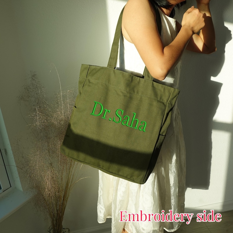 Pode incluir: Bolsa tote verde-oliva com o texto bordado "Dr. Saha" em verde. A bolsa tem al&ccedil;as longas e formato retangular. As palavras "Embroidery side" est&atilde;o escritas em rosa na parte inferior da imagem.
