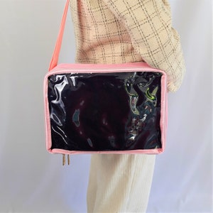 K&ouml;nnte beinhalten: Eine rosa Stoffaufbewahrungstasche mit einem transparenten Plastikfenster. Die Tasche hat einen Schultergurt und wird mit einem Rei&szlig;verschluss geschlossen.