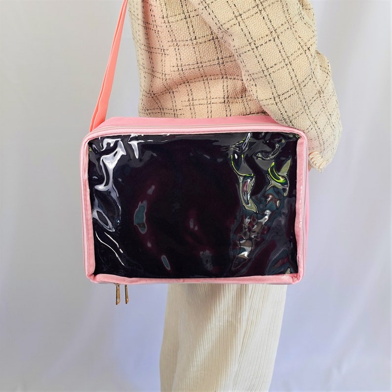 K&ouml;nnte beinhalten: Eine rosa Stoffaufbewahrungstasche mit einem transparenten Plastikfenster. Die Tasche hat einen Schultergurt und wird mit einem Rei&szlig;verschluss geschlossen.