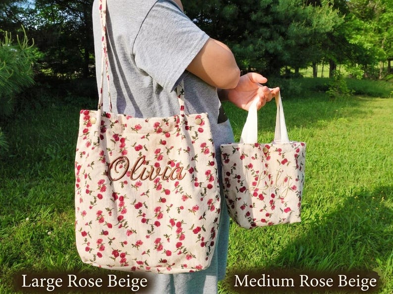 May include: Two beige floral tote bags, one large and one medium, with red rose patterns. The large bag has a shoulder strap and the name "Olivia" embroidered on it. The medium bag has a handle and a name embroidered on it.