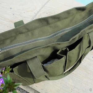 May include: Olive green canvas tote bag with a zippered top and multiple exterior pockets. The bag has sturdy handles and a neutral-colored lining. The bag is sitting on a light-colored surface.