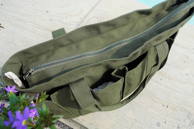 May include: Olive green canvas tote bag with a zippered top and multiple exterior pockets. The bag has sturdy handles and a neutral-colored lining. The bag is sitting on a light-colored surface.