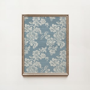 May include: Framed floral print featuring a blue background with white floral patterns. The artwork is enclosed in a rectangular, light brown wooden frame. The floral design includes detailed flowers and leaves.