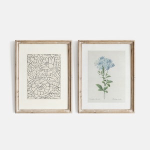 Puede incluir: Dos impresiones enmarcadas, una con un patr&oacute;n floral en blanco y negro y la otra con una pintura de acuarela de flores azules. Ambos marcos son de madera clara con un passe-partout blanco.