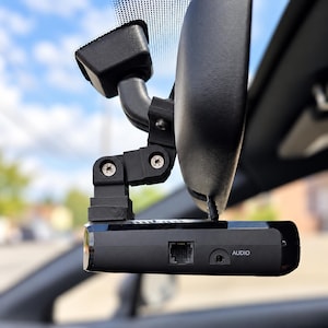 May include: A black dash cam mounted to a rearview mirror with a black plastic adjustable bracket. The device features a rectangular body with a visible Ethernet-style port and a circular audio jack labeled with the word AUDIO in white text.