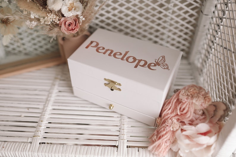 K&ouml;nnte beinhalten: Wei&szlig;e Holz-Musikbox mit goldenem Verschluss und einer Schublade. Die Box tr&auml;gt den Namen "Penelope" und ein Schmetterlingsdesign auf dem Deckel.
