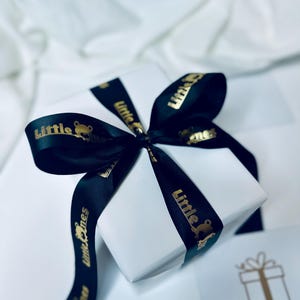 K&ouml;nnte beinhalten: Eine wei&szlig;e Geschenkbox mit einem schwarzen Band, das zu einer Schleife gebunden ist. Das Band tr&auml;gt die Aufschrift "Little Ones" in Gold.