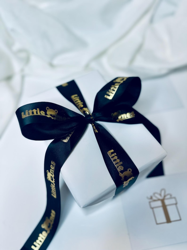 K&ouml;nnte beinhalten: Eine wei&szlig;e Geschenkbox mit einem schwarzen Band, das zu einer Schleife gebunden ist. Das Band tr&auml;gt die Aufschrift "Little Ones" in Gold.