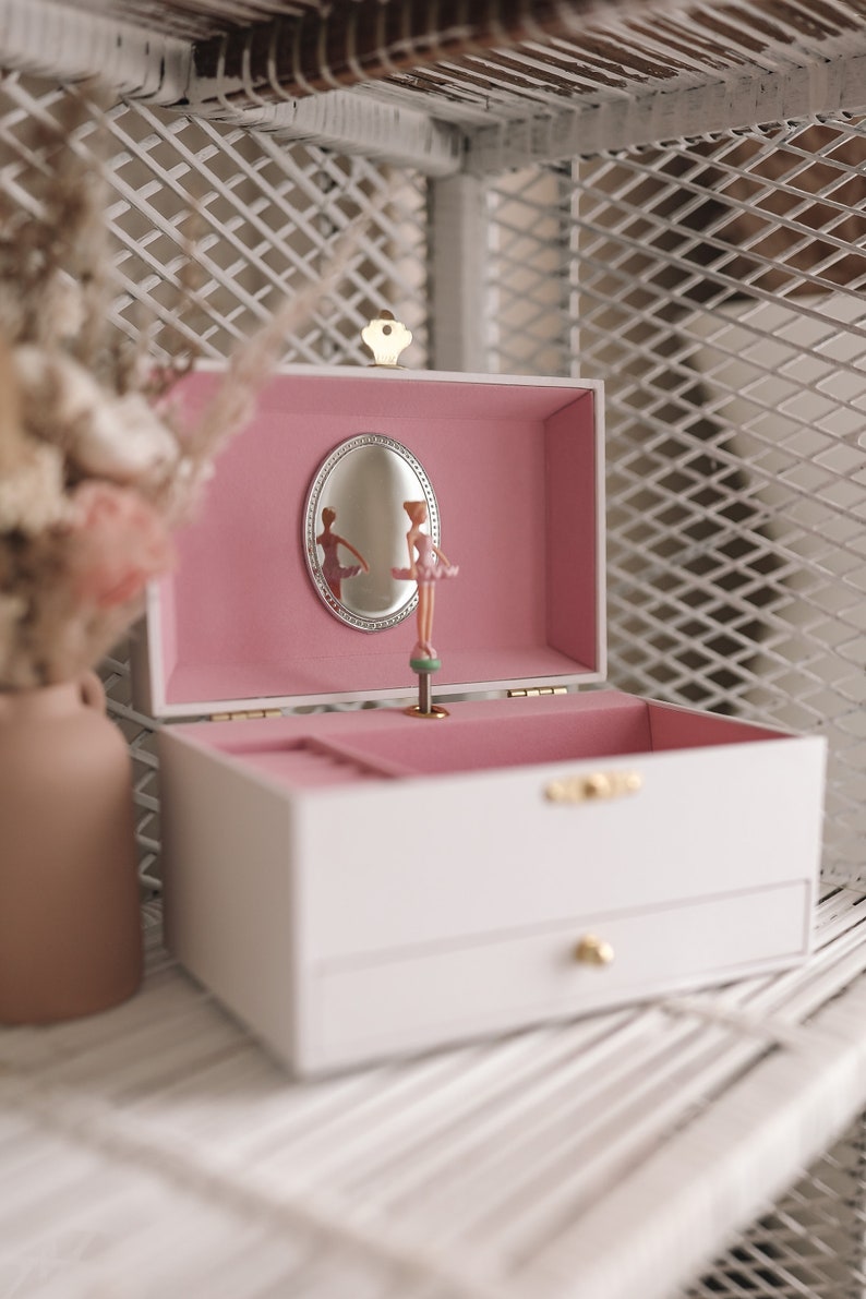 K&ouml;nnte beinhalten: Wei&szlig;e Spieluhr mit rosa Innenraum und einer Ballerina-Figur im Inneren. Die Box hat eine kleine Schublade und ein goldenes Schl&uuml;sselloch.