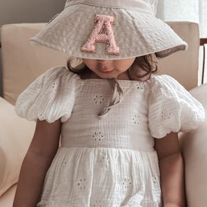 May include: A young child wearing a white dress with a floral pattern and a light grey sun hat with a pink letter 'A' on the front.