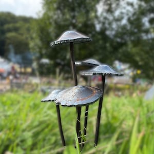 Könnte beinhalten: Ein Satz von vier Pilzskulpturen aus Metall mit einer blau-grauen Oberfläche. Die Skulpturen sind in einer Gruppe angeordnet, wobei der größte Pilz in der Mitte und die kleineren Pilze ihn umgeben. Die Pilze sind aus Metall und haben ein rustikales, verwittertes Aussehen.