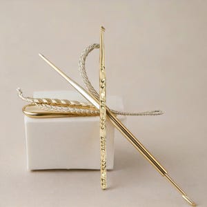 May include: Two gold crochet hooks with a twisted design. The hooks are on a white surface with a white and silver twisted cord.
