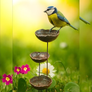 May include: A tiered metal bird feeder featuring three small, dark, bowl-shaped basins stacked vertically on thin metal rods. A small blue, yellow, and white Eurasian blue tit bird is perched on the rim of the top basin, looking to the left.