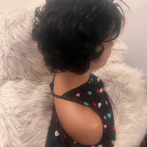 May include: A mannequin head with short, dark, curly hair. The mannequin is wearing a black top with a pattern of colorful hearts. The background is a fluffy, white, faux fur material.