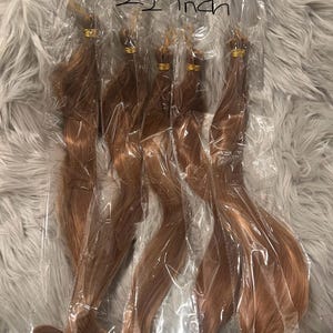 May include: Five bundles of wavy, auburn-colored hair extensions, each sealed in a clear plastic bag. The hair is tied with a gold band. The text "Color=M30/27" and "22 inch" is written on the packaging.