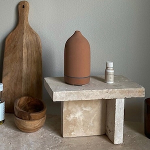 May include: A terracotta essential oil diffuser on a beige stone pedestal with a small bottle of essential oil, a wooden cutting board, a cookbook titled "Half Baked Harvest super simple", and a bottle of olive oil.