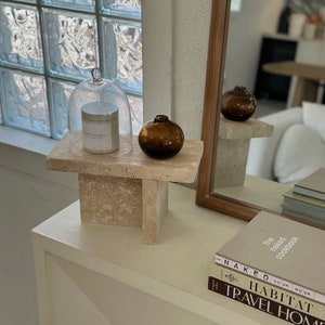 May include: A white candle with a glass cloche, a brown glass vase, and a stack of books with the title "The Naked Cookbook" on top. The items are displayed on a small, square, beige stone pedestal.