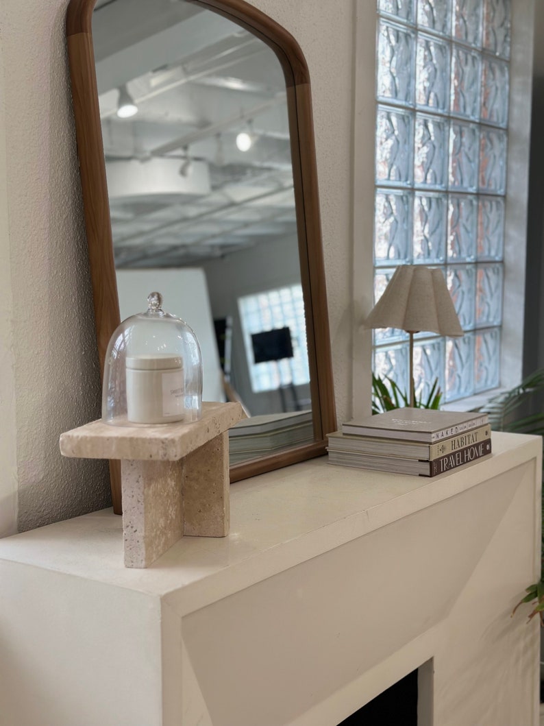 May include: A white mantelpiece with a white candle in a glass cloche on a stone pedestal. A lamp with a white shade sits on a stack of books with the titles "Maker", "Habitat", and "Travel Home".