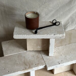 May include: A brown leather candle jar with a white candle inside, a black candle snuffer, and a stack of beige stone tiles.