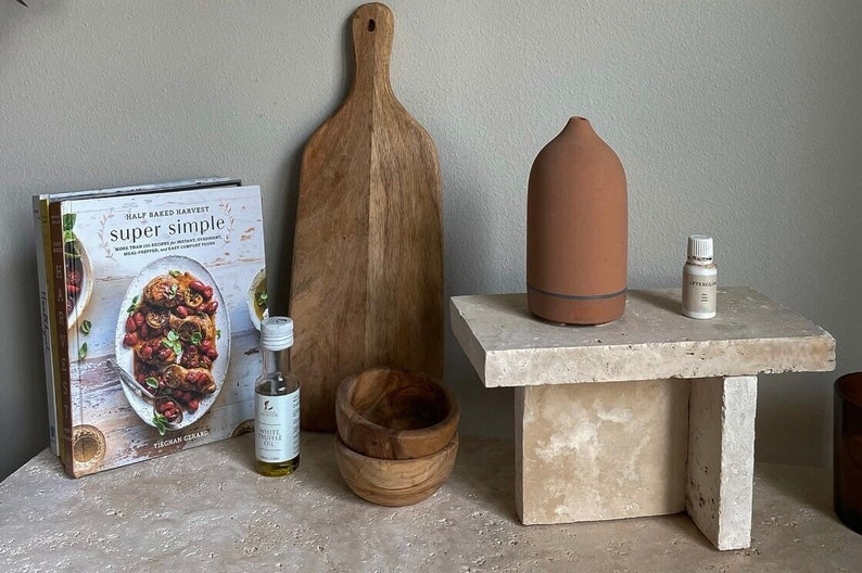 May include: A terracotta essential oil diffuser on a beige stone pedestal with a small bottle of essential oil, a wooden cutting board, a cookbook titled "Half Baked Harvest super simple", and a bottle of olive oil.