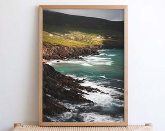 Peut inclure: Une photographie de paysage encadrée montrant une côte accidentée avec des falaises rocheuses sombres rencontrant des vagues turquoise. Au-dessus des falaises, une colline vert vif s'élève, parsemée de petites maisons blanches.