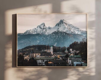 May include: A square framed wall art print featuring a landscape photograph of a snow-capped mountain range towering over a town with traditional buildings, a church steeple, and dense forest under a soft, overcast sky in muted tones.