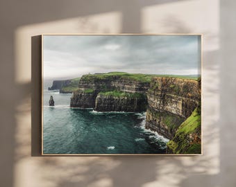 Peut inclure: Une impression d'art mural rectangulaire présentant une photographie de paysage des falaises de Moher. L'image montre des formations rocheuses sombres et stratifiées surmontées d'herbe verte vive, rencontrant l'océan bleu sous un ciel nuageux.