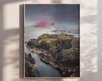 May include: A framed wall art print featuring a white lighthouse perched on a rugged, green-covered rocky cliffside overlooking dark water. The sky above displays a dramatic blend of grey clouds with a soft pink sunset glow on the horizon.